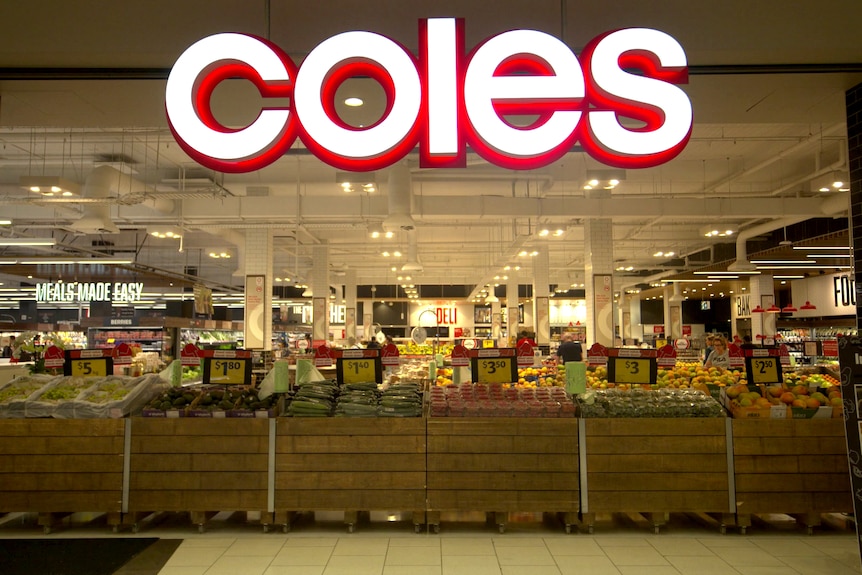 The front of a Coles supermarket. Underneath a large sign are displays of fruit and vegetables.