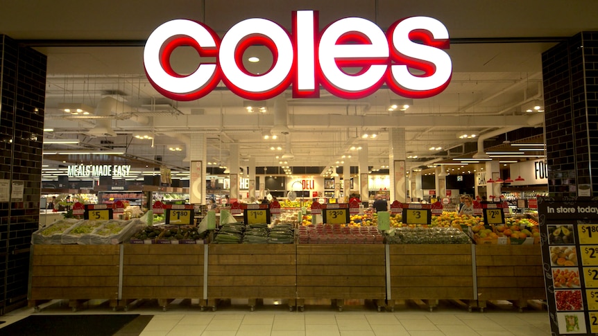 The front of a Coles supermarket. Underneath a large sign are displays of fruit and vegetables.