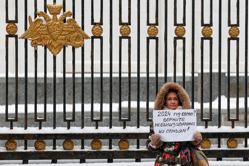 A woman with a large coat on holds a sign and stands outside a large fence as snow falls 