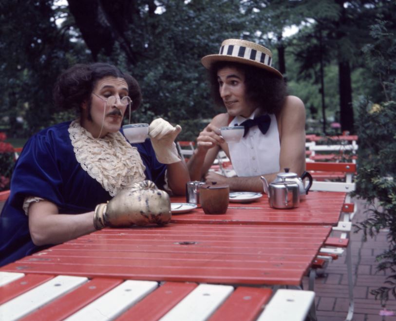 Two characters sit at a table sipping tea.