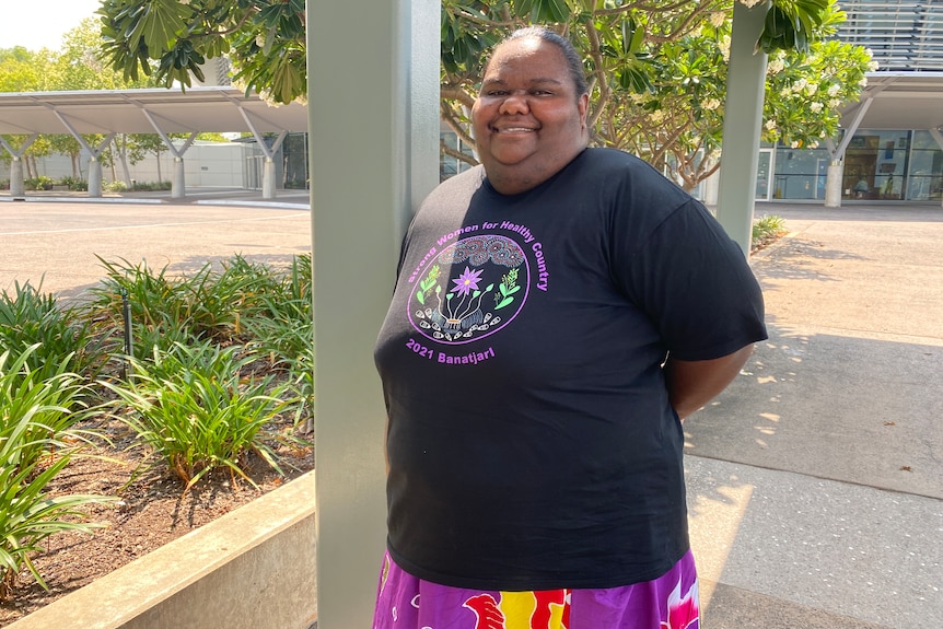 An Aboriginal woman wearing a Strong Women for Healthy Country shirt smiles with her hands behind her back.