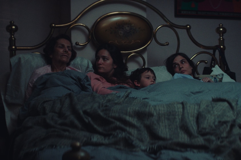 An older woman with nasal oxygen cannula sleeps propped up in bed next to two women (and a child) who look at her with concern.