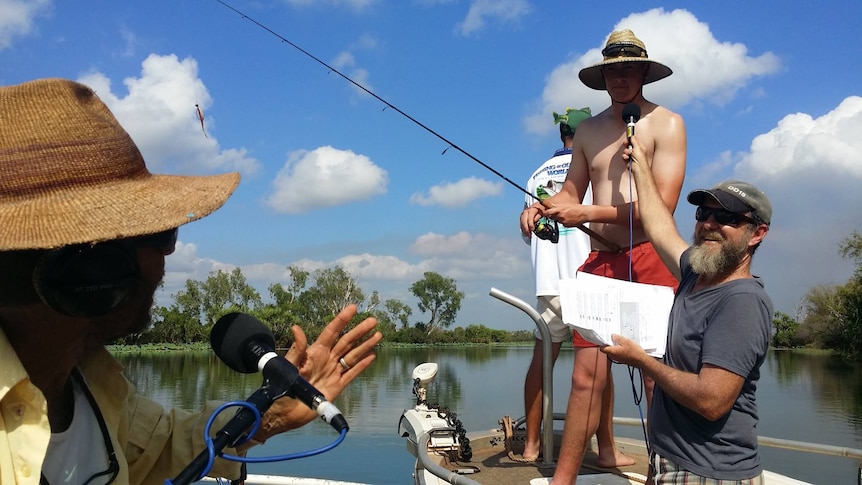 Man stands on boat with fishing rod while being interviewed for radio