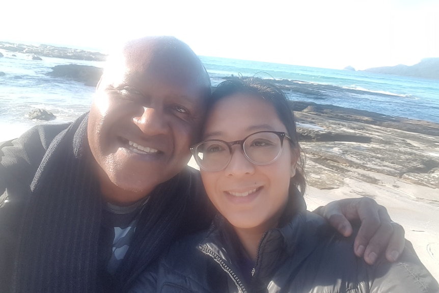 A selfie of a man and woman with their arms around one another's shoulders, smiling, at the beach.