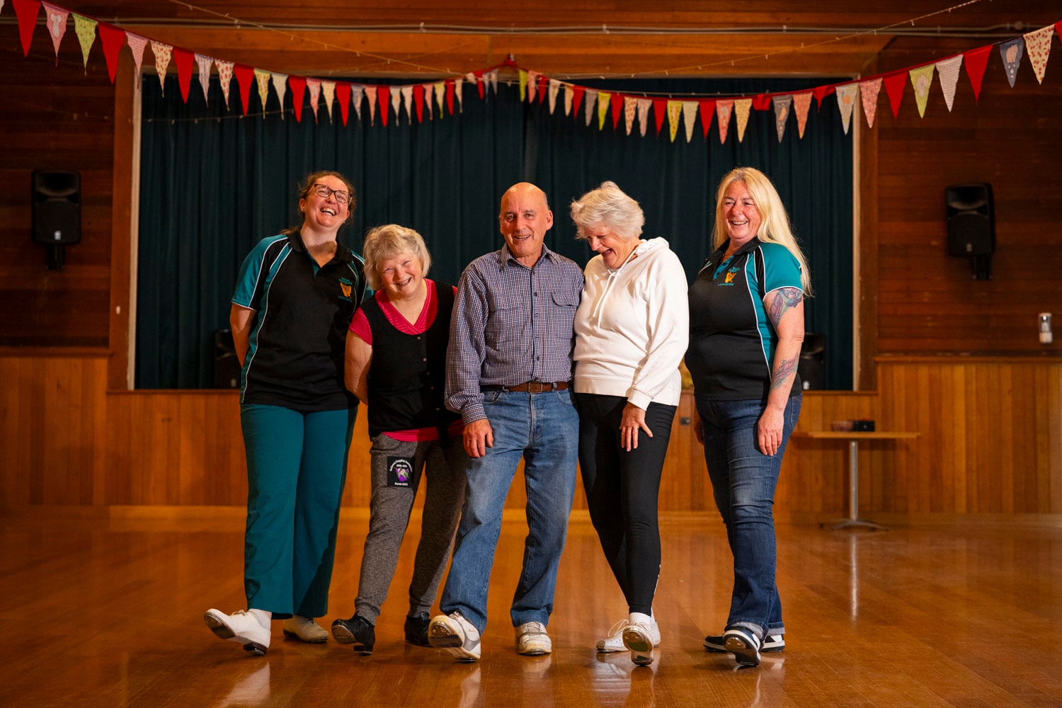 A group of 5 dancers in tap shoes, in a=a wooden hall surrounded by little flags