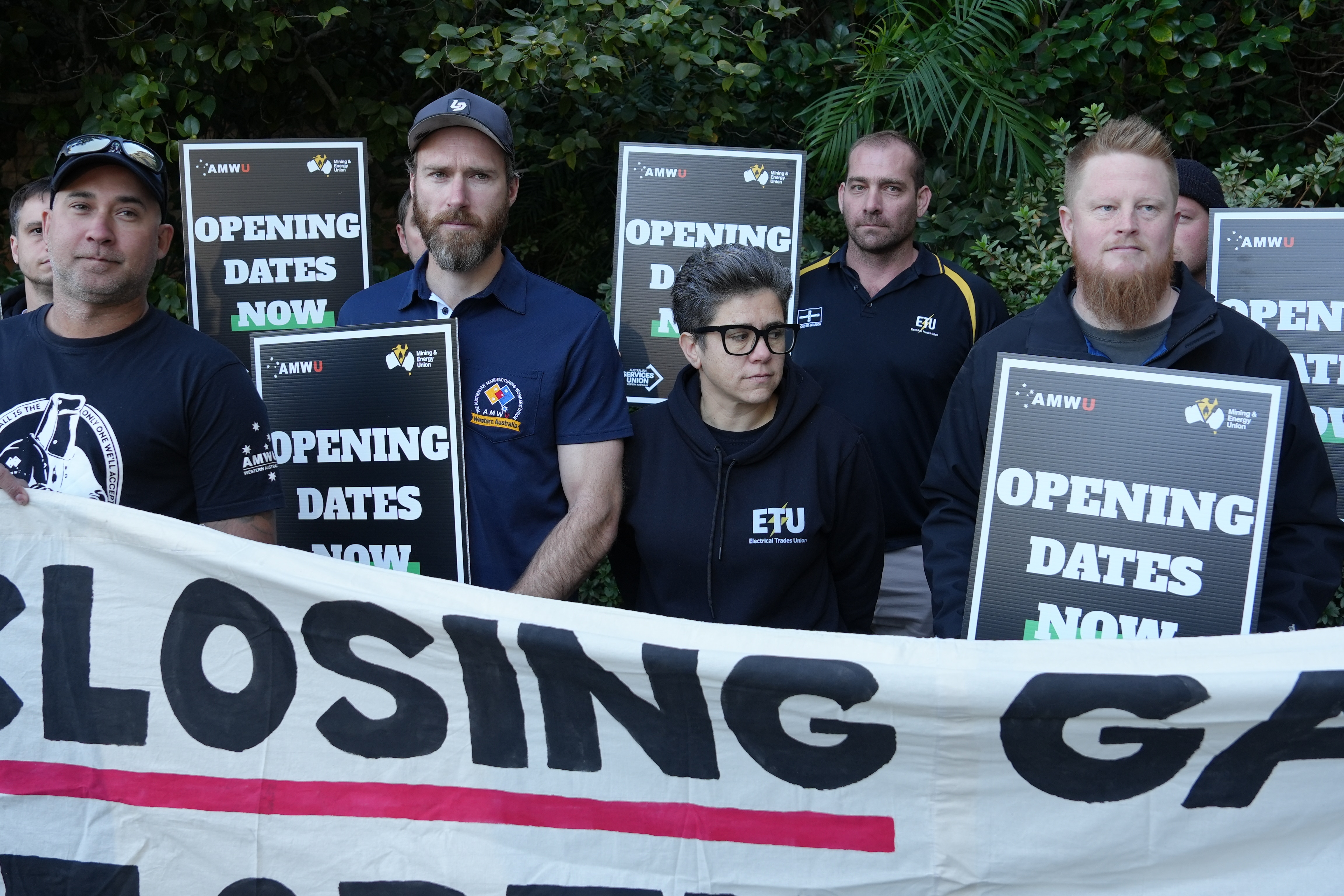 Six protesters standing holding black signs behind a white banner which read 'opening dates now'