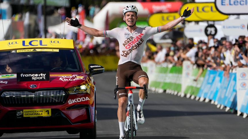 A cyclist wears a beaming smile as he spreads his arms wide in triumph crossing the finish line in a Tour de France stage. 