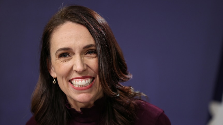 Jacinda Ardern, wearing mauve top and lipstick, grins widely in a close-up shot against a dark blue background