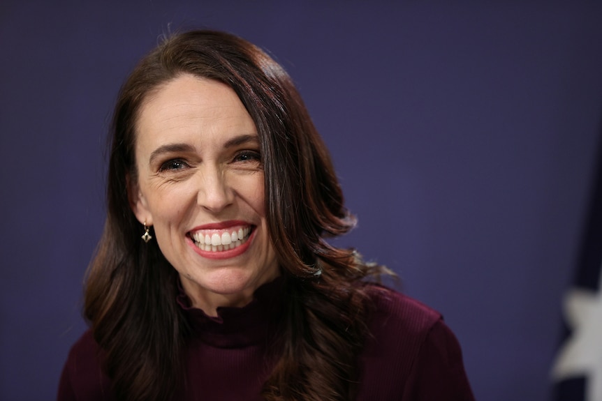 Jacinda Ardern, wearing mauve top and lipstick, grins widely in a close-up shot against a dark blue background