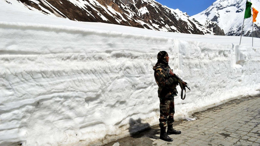 An Indian army soldier stands guard on a mountain road with his back to a wall of snow taller than him.