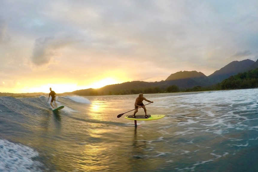 A surfer rides a wave behind another surfer on a different type of board, using a paddle.