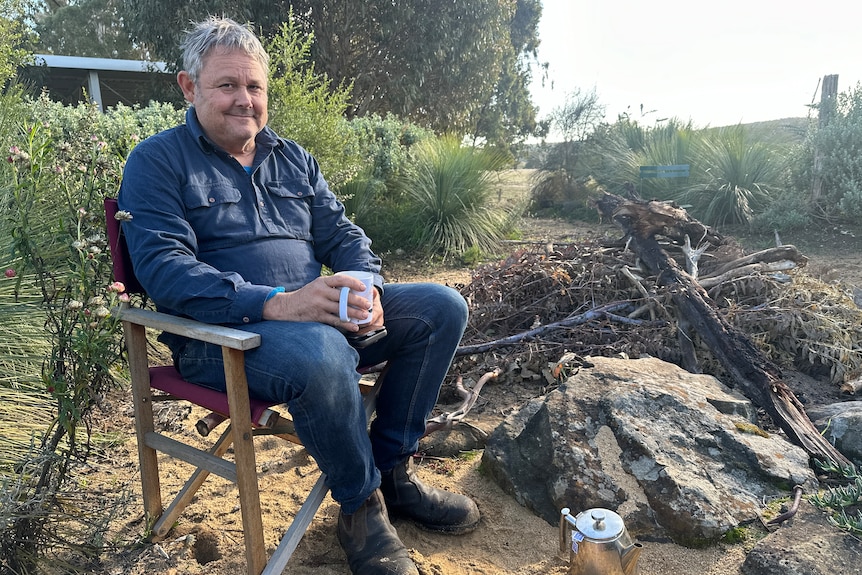 A man sitting in a wooden fold-out camping chair with a tea cup in his hands and a pot of tea at his feet, sitting outside.