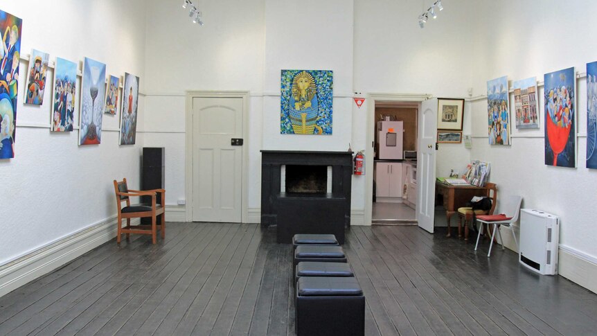 A large hall with white walls, black stools in the centre of the room and paintings on the walls