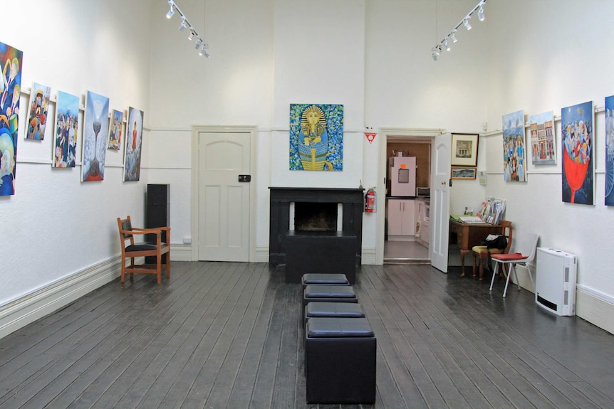 A large hall with white walls, black stools in the centre of the room and paintings on the walls