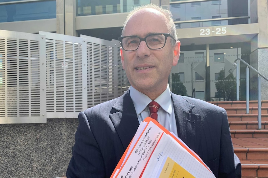 A man in glasses holding folders stands in front of court building