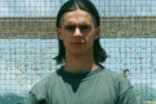 A young man with dark, shoulder length hair stands against a prison fence.