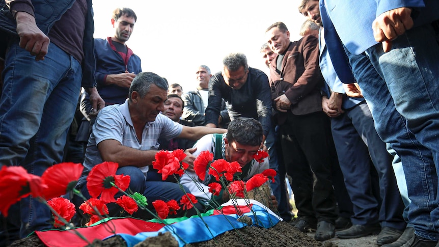 A weeping man crouches over a grave topped with poppies surrounded by a group of men.