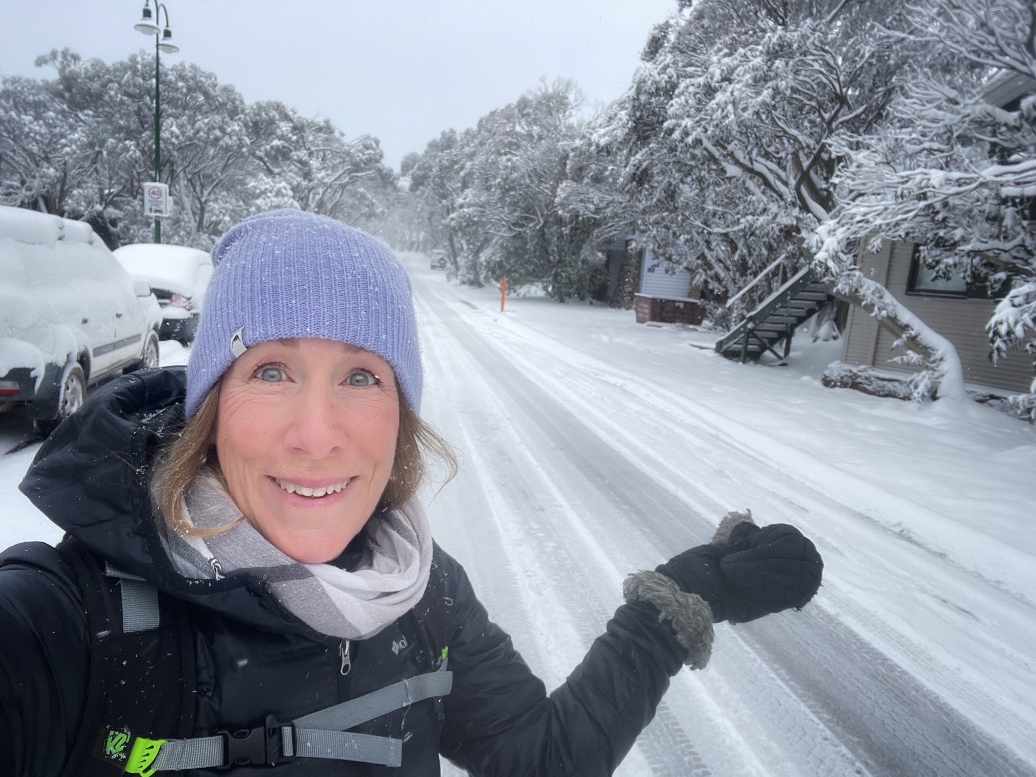 A smiling woman in a beanie stands on the side of a snow-covered road.