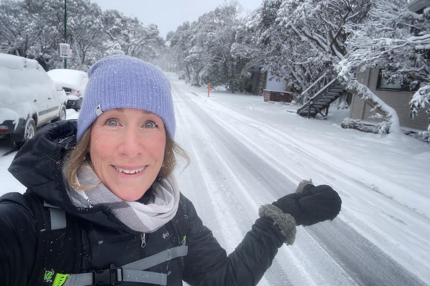 A smiling woman in a beanie stands on the side of a snow-covered road.