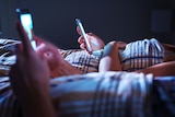 Two people lie in bed, using their separate smartphones.
