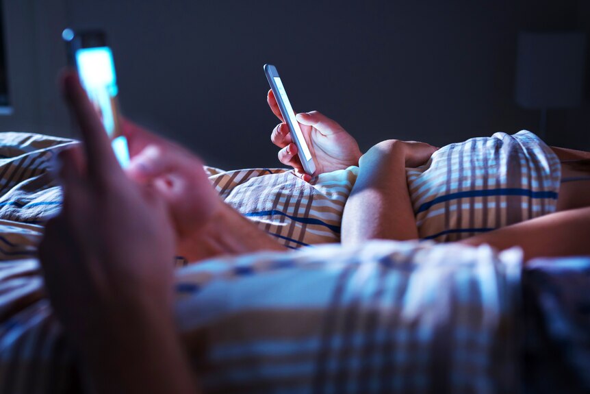 Two people lie in bed, using their separate smartphones.