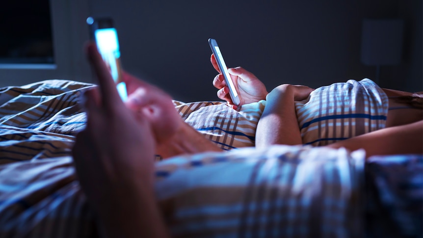 Two people lie in bed, using their separate smartphones.