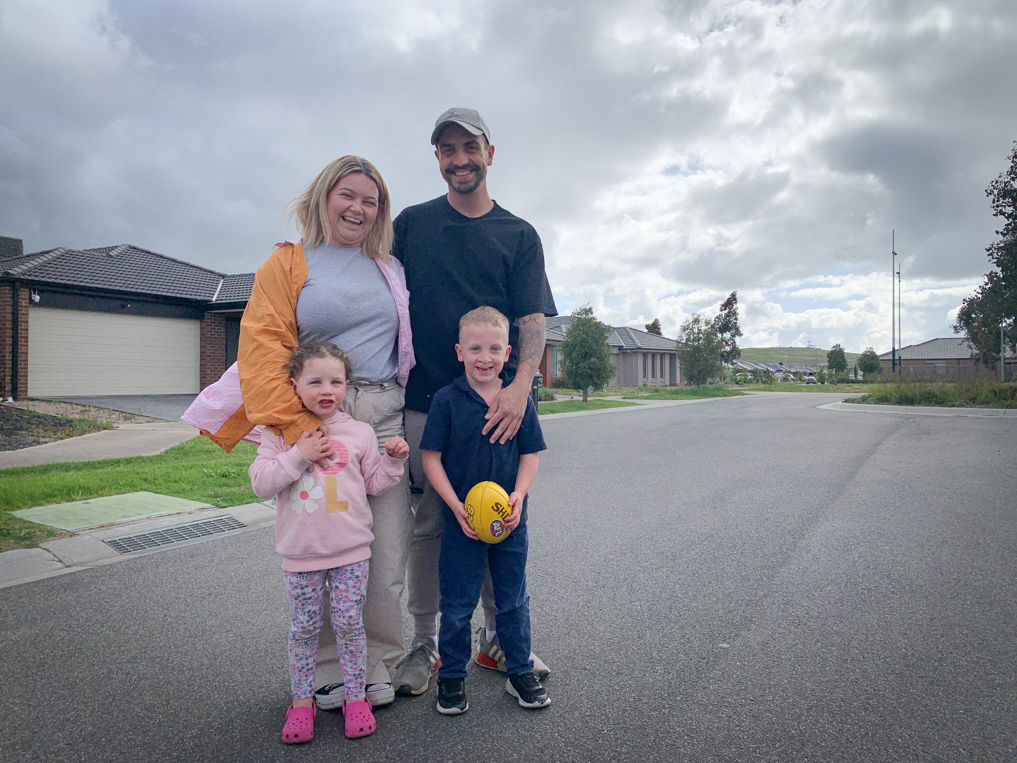 Tristan Wilson smiling in a portrait with his wife and two young children, standing on the street outside their home.