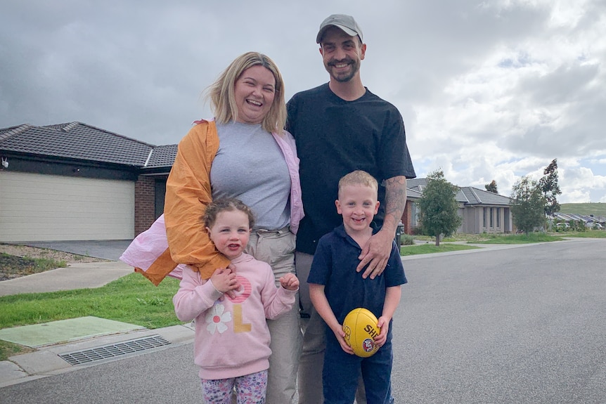 Tristan Wilson smiling in a portrait with his wife and two young children, standing on the street outside their home.