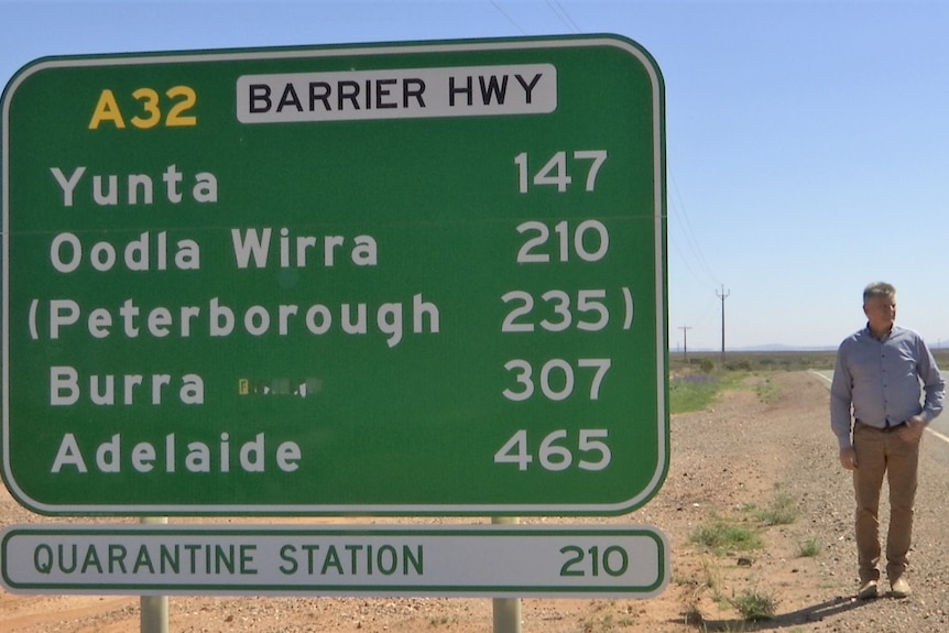 A man stands beside a huge sign on a highway showing distances to destinations.