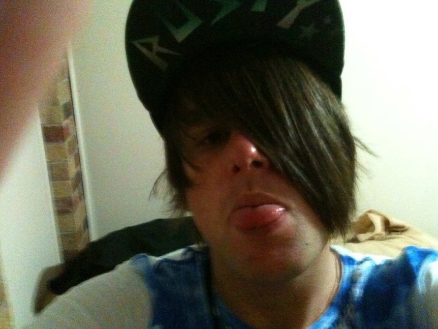 A young man with dark hair wearing a baseball cap sticks his tongue out as he poses for a selfie.