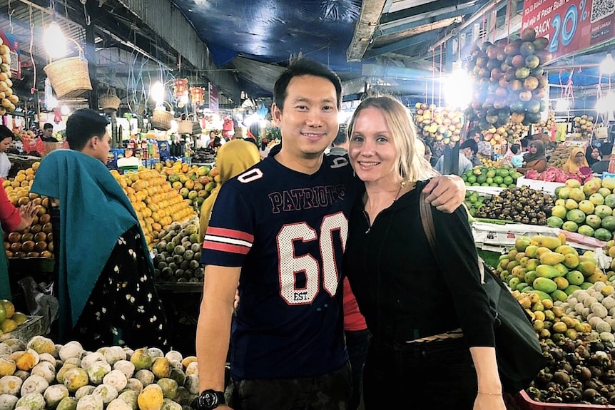 Man with his arm around woman's shoulder poses for a photo at a fruit market.