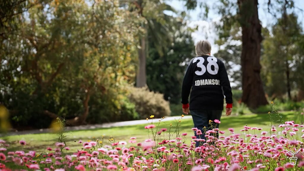 Jane walking through a garden filled with pink flowers wearing a jumper, 38 Edmanson.
