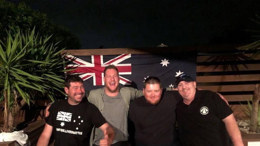 Four men in black t-shirts sit in front of an Australian flag