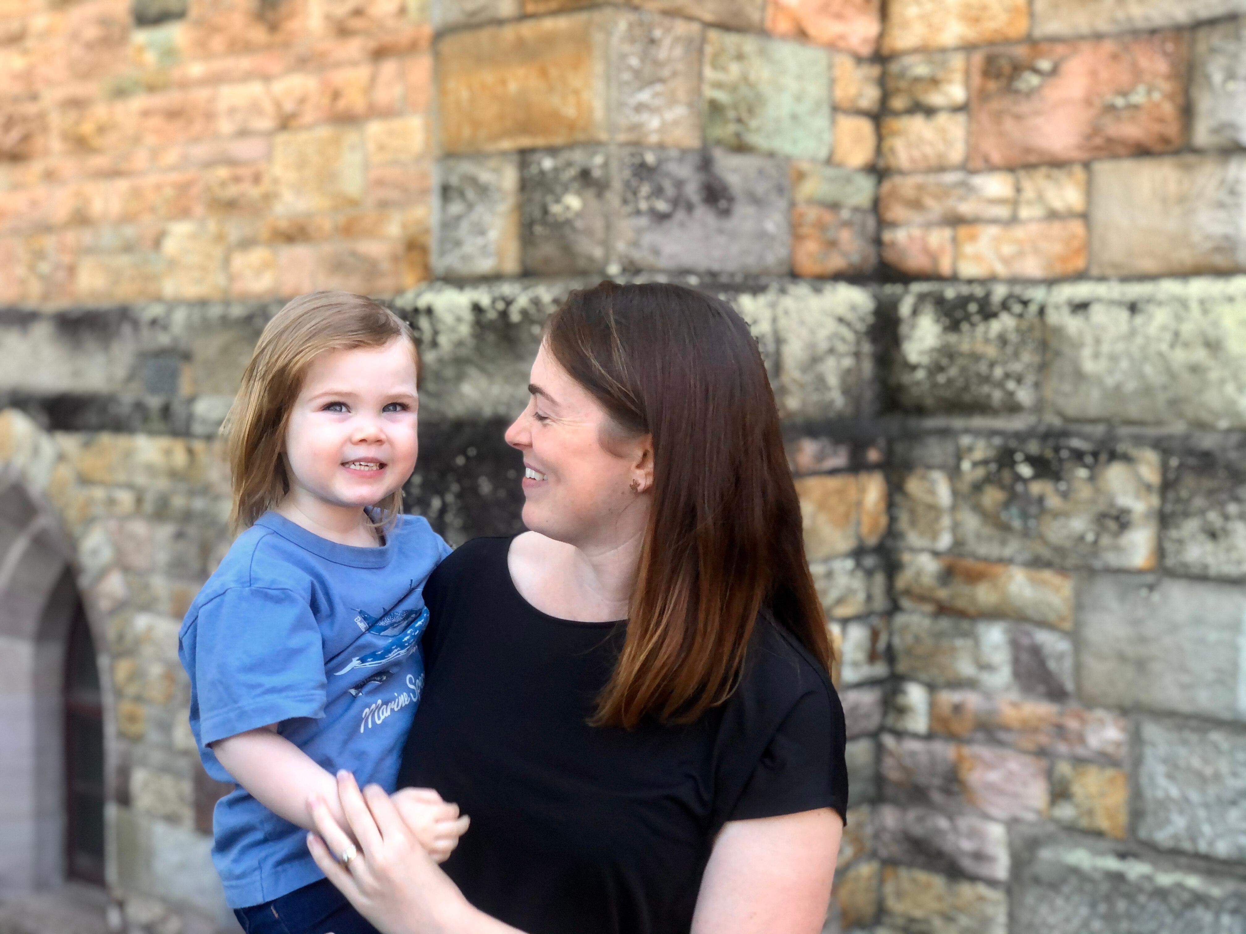 An image of Sarah Muchall holding her daughter Eleanor and smiling in her direction in front of a brick church building. 