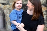 An image of Sarah Muchall holding her daughter Eleanor and smiling in her direction in front of a brick church building. 