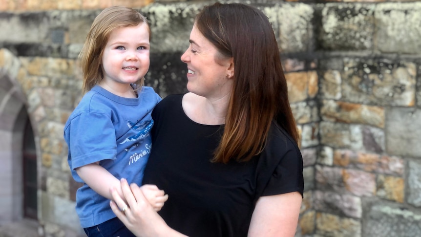 An image of Sarah Muchall holding her daughter Eleanor and smiling in her direction in front of a brick church building. 