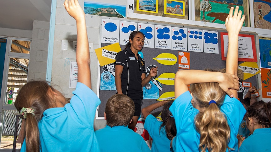 A teacher in a classroom in front of students, two with their hands raised.