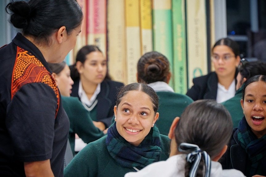 A female student smiles up at the woman talking to her, surrounded by her classmates wearing green, blue & white