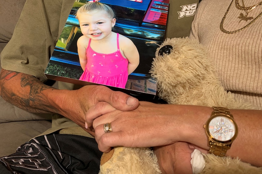 Close up image of parents holding hands with young daughter's photo and stuffed toy.