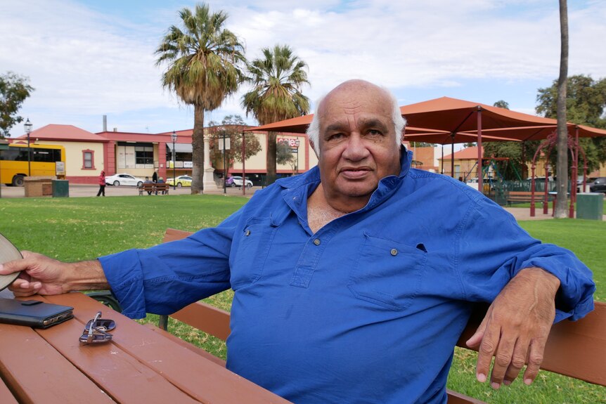 An Indigenous man sitting on a bench in a park wearing a blue shirt