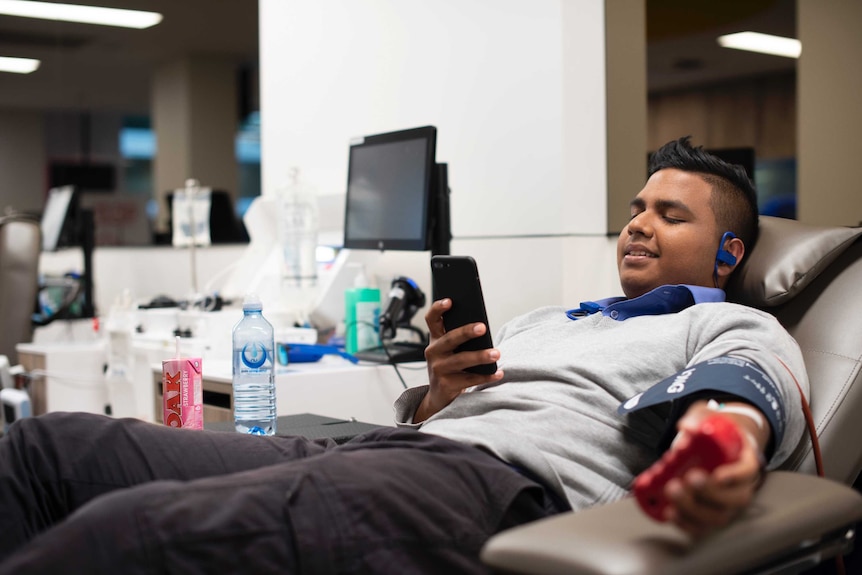 Blood donor reclining in chair and looking at phone while blood is being taken