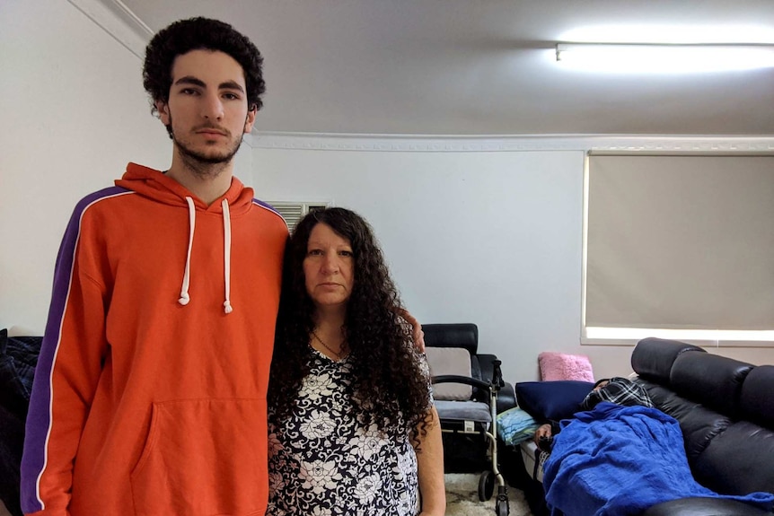 A young man stands next to his mum with his hand around her shoulder in their lounge room