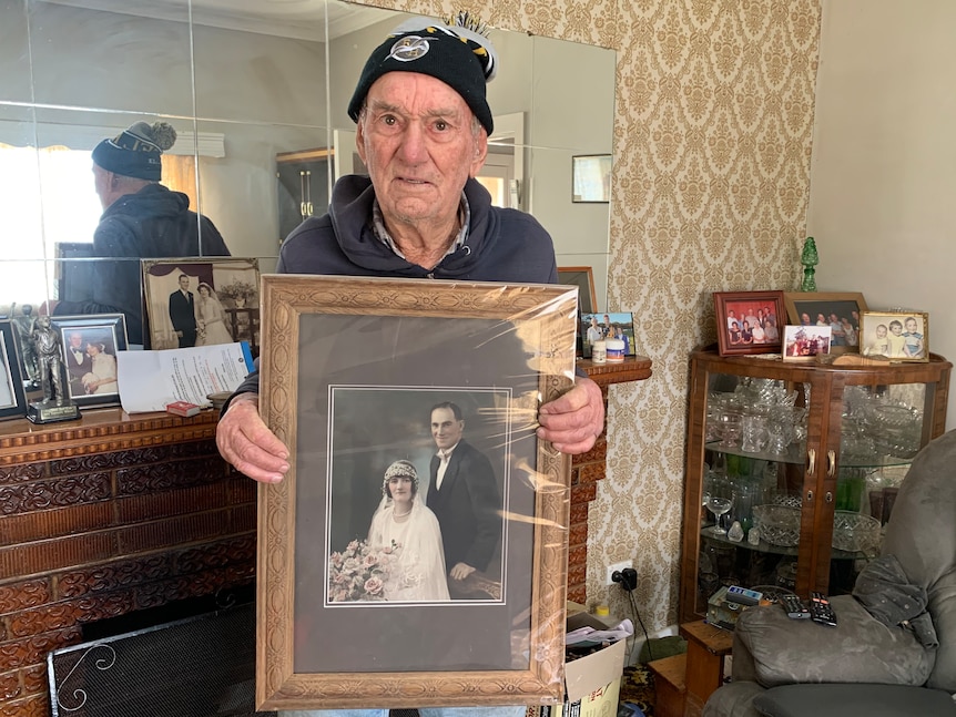 A man stands in a living room in a mirror on the wall holding a photo of a couple in wedding garb. A drinks cabinet in corner