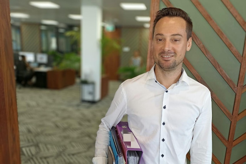 A man wearing a white button-up top holding files in an open-plan office 