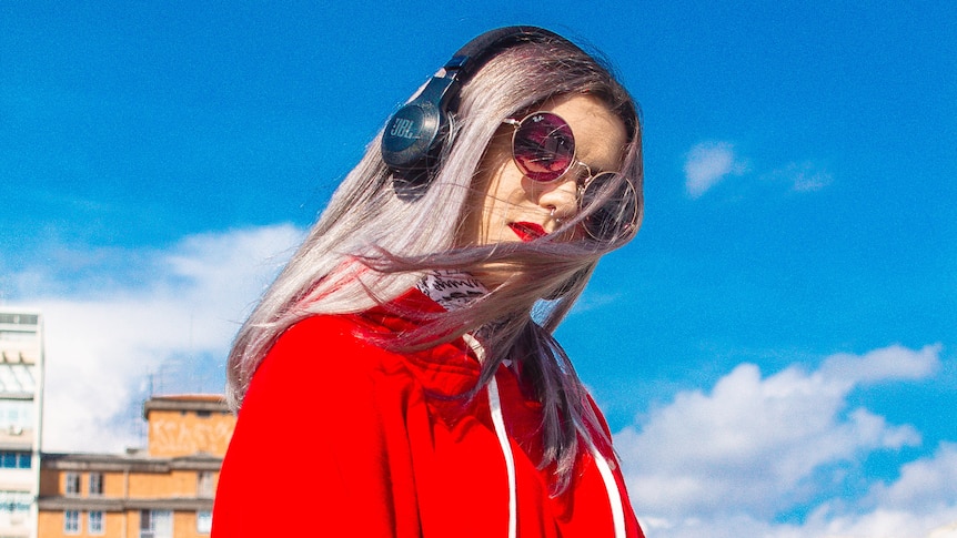 A woman with should-length hair, red lipstick, red jacket and grey headphones looks down towards the camera.