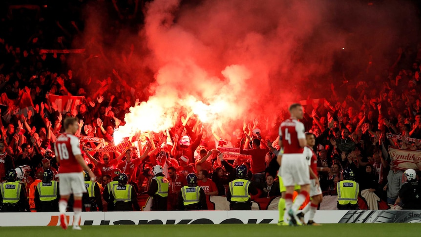 Arsenal players look on as FC Koln supporters light flares
