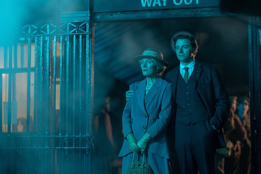 Older white woman in suit jacket and skirt with gloves and hat stands beside white man in three-piece suit on blue-lit platform.