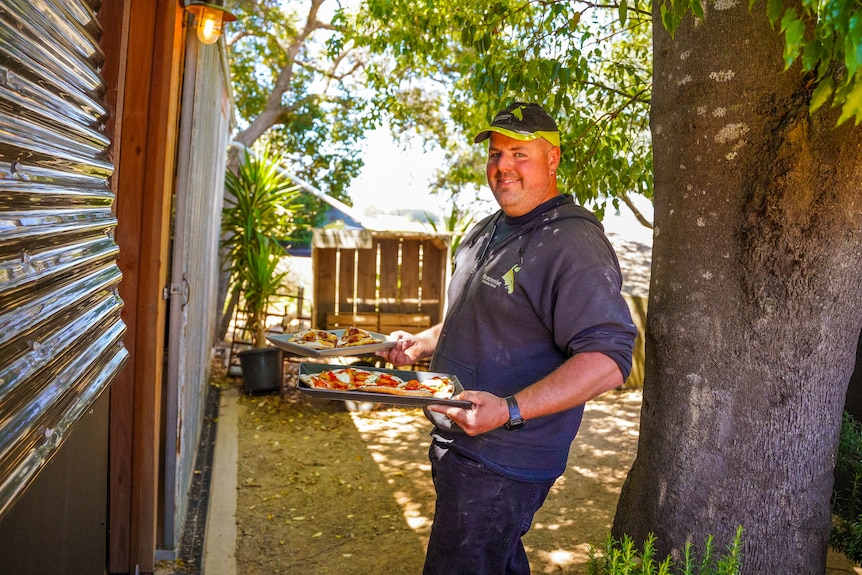 A man in a black hooded jumper and cap carries a tray of pizza smiling.