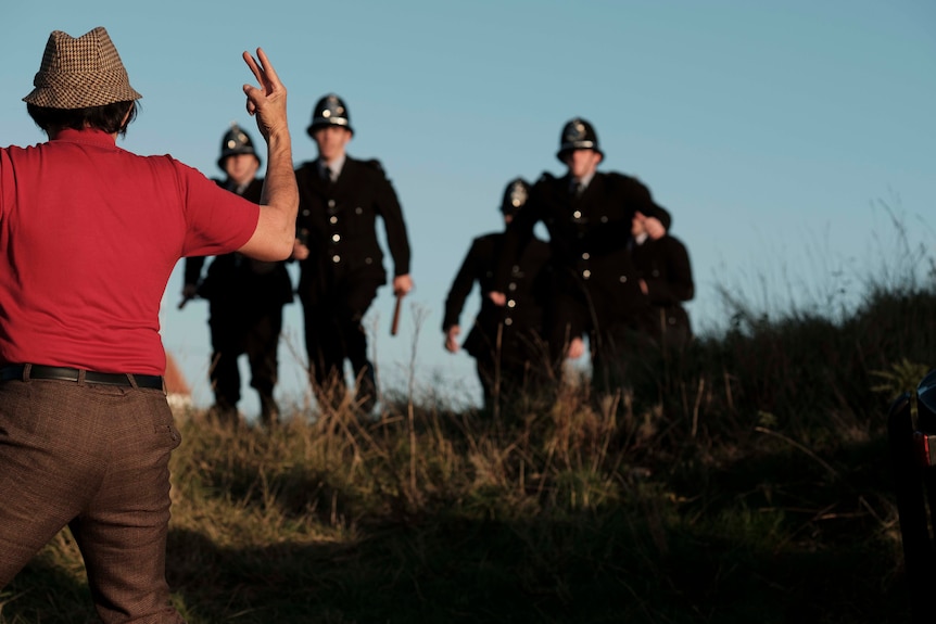 An older man, his back to the camera, makes an "Up yours" hand gesture at a group of policemen, chasing him on a golf course