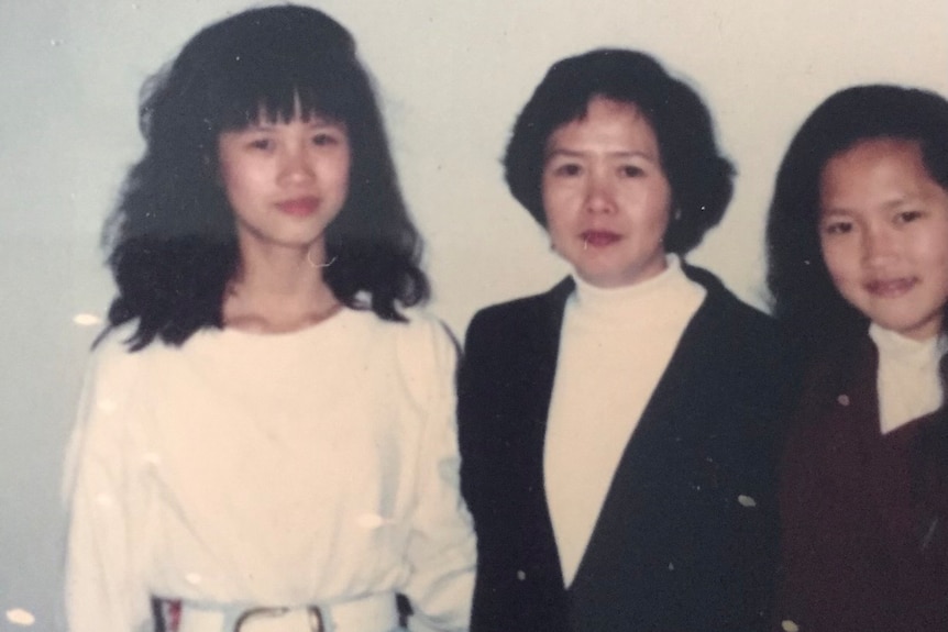 Girl in white top and blue jeans looks to camera next to two women.